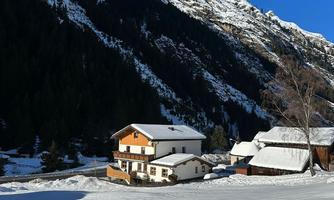 A picturesque winter landscape with snow-covered hills and a clear blue sky. In the foreground, there are cozy wooden and stone farmhouses.