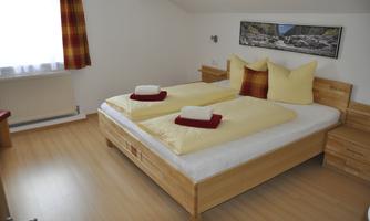 A cozy bedroom with a double bed and light wooden furniture. The bedding is yellow with red accents, and there is a large window that lets in natural light.