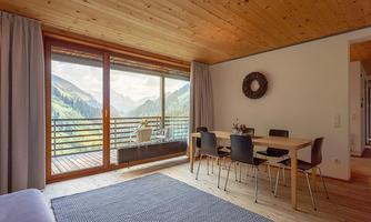 A modern living room with a large window that offers a view of the mountains. The room is minimally furnished with a dining table and chairs.