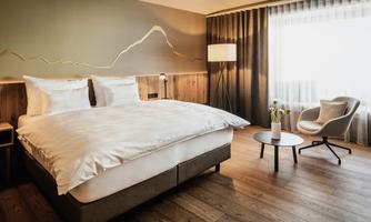 A modern hotel room with a large bed and simple furnishings. The room has wooden flooring and a window with plenty of daylight.