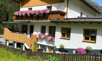 A picturesque house in the mountains with a balcony full of flowers. The garden is green and inviting, surrounded by beautiful nature.