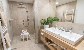 A modern bathroom with tiles in neutral colors and a glass shower. On the sink countertop, there is a small plant and a welcome sign.