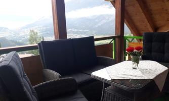 A cozy balcony with rattan furniture and a table on which a flower vase stands. In the background, mountains and a cloudy sky can be seen.