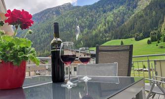 A balcony with a beautiful view of the mountains and a green meadow. Two wine glasses and a bottle of wine are on the table, next to a blooming flower.