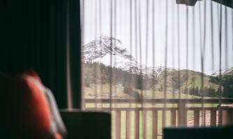 A view through a curtain onto a green landscape with snow-covered mountains in the background. The scene conveys a calm and relaxing atmosphere.