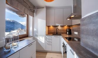 A modern kitchen with white cabinets and wooden details. Large windows offer a view of the mountains and provide plenty of daylight.