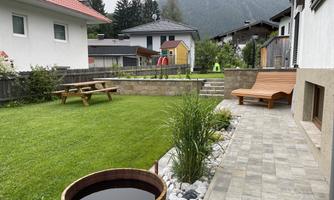 A beautiful garden with grass, a wooden bench, and a round wooden basin. In the background, houses and mountains can be seen.