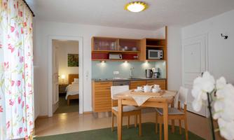 A modern kitchen with a dining area and bright furniture. In the background, a bedroom can be seen.