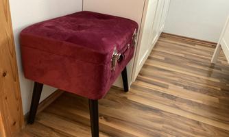 An elegant stool in dark red with four black legs. It stands in a corner of a room on a wooden floor.