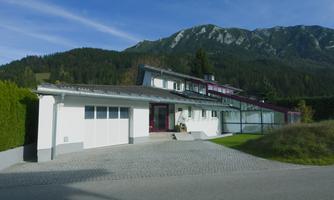 A modern house in a rural setting, surrounded by mountains and trees. The garden is well-maintained and the driveway is paved with cobblestones.