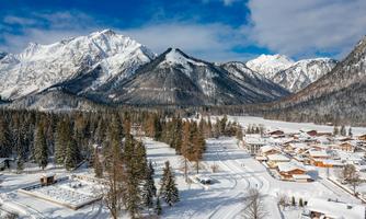 A picturesque winter landscape with snow-covered mountains and an idyllic village. The trees are covered in snow and the sky is partly cloudy.