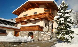 A charming chalet in Tyrolean style with wood decorations and a snow-covered garden. The clear blue sky creates an inviting winter atmosphere.