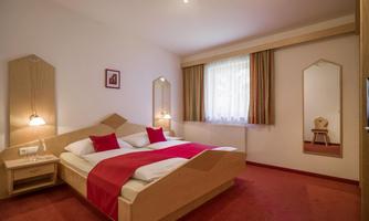A cozy hotel room with a double bed and red cushions. The walls are bright, and there is a large mirror as well as a window with curtains.
