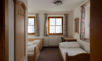 A simple bedroom with two single beds and a table. The windows are decorated with curtains, and there are light walls and wooden details.