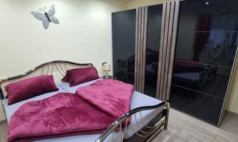 A modern bedroom with a large bed and red pillows. In the background, there is a black wardrobe and soft lighting.