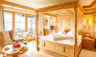 A cozy room with a wooden four-poster bed and warm colors. Large windows provide plenty of daylight and offer a view outside.