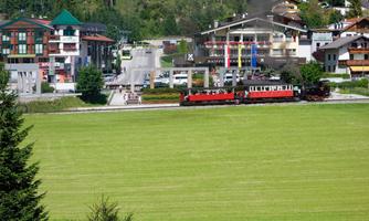 A red steam locomotive travels through a green meadow. In the background, cozy buildings and trees can be seen.