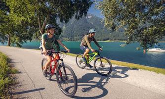 Two cyclists ride along a picturesque lake. In the background, mountains stretch under a clear sky.