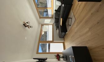 A modern living room with a comfortable black sofa and large windows. The space has a warm wooden floor and is bright and inviting.