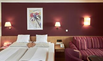A modern hotel room with two beds and a cozy seating area. The walls are painted in a deep violet, and decorative lighting creates an inviting atmosphere.
