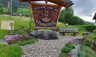 A wooden sign welcomes visitors to Kolsassberg. Surrounded by colorful flowers and a picturesque landscape.