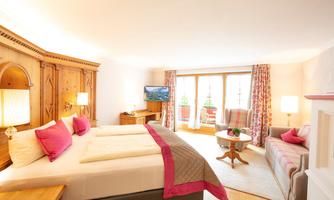 A bright room with two beds, decorated walls, and a beautiful view of the balcony. There is a table, a television, and cozy seating.