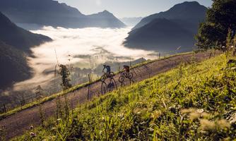 Two cyclists ride along a path through a beautiful mountainous landscape. The fog lies gently in the valleys, and the sun shines on the surrounding mountains.