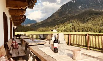 A cozy terrace with wooden furniture and a view of the mountains. In the background, green forests and a blue sky can be seen.