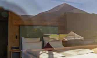 A cozy bedroom with a large window reflecting a mountain landscape. The furniture is made of wood, and the bed linens look inviting.