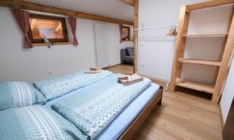 A cozy bedroom with two beds and blue bedcovers. There is a shelf and windows with red curtains that provide plenty of daylight.