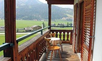 A cozy balcony with a wooden railing and a table with a chair. In the background, green meadows and mountains can be seen.