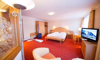 A cozy hotel room with two beds, a television, and a comfortable armchair. The room is bright and friendly with a beautiful carpet.