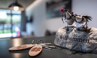 An inviting reception scene with a stone bearing the word "Welcome," and keychains next to a decorative bird. In the background, modern furniture and natural light are visible.