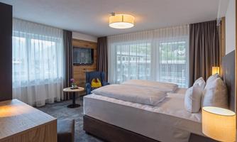 A modern hotel room with a large bed and a cozy armchair. The large windows let in plenty of light and offer a view of the surroundings.