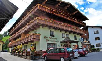 A traditional Austrian inn with balcony decorations and blooming flowers. The surroundings feature a quiet street and several parking spaces.