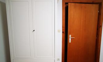 An elegant white wardrobe with two drawers is in a room. In the background, a wooden door is visible.