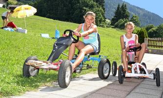 Two girls are riding go-karts on a paved path. In the background, there are fields and trees.