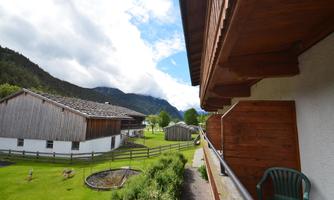 A view from a balcony over a green landscape with traditional wooden houses and mountains in the background. The sky is partly cloudy and it looks calm and inviting.