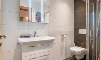 A modern bathroom with a shower, a sink, and a toilet. The walls are designed in light and dark colors, and there is a mirror with lighting.