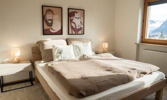 A cozy bedroom with a large double bed and soft, neutral colors. Two animal pictures hang on the wall, and the window offers a view of the mountains.