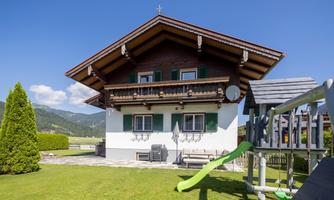 A traditional two-story house in alpine style. In the foreground, you can see a playground with a slide and a green meadow.
