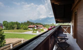 A beautiful balcony with a view of green meadows and mountains. On the balcony, there are two chairs and flower boxes.