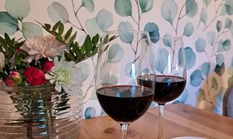 Two wine glasses filled with red wine are placed on a wooden table, accompanied by a flower vase and a bowl of snacks. In the background, there is a wall with gentle green leaves.