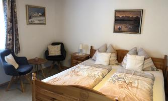 A cozy bedroom with a large bed and beige-white bedcovers. There is a small table and an armchair in the corner, as well as appealing wall pictures.