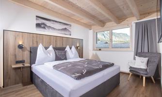 A modern bedroom with a comfortable double bed and a wooden ceiling. Large windows provide plenty of daylight.