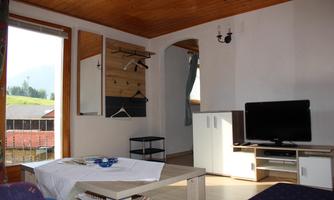 A cozy living room with wooden floor and light walls. There is a table, a television, and a window view of the landscape.