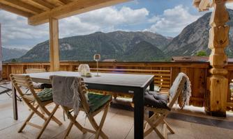 A cozy terrace with a table and chairs, surrounded by stunning mountains. The sky is clear and the landscape is green and inviting.