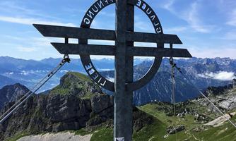 A prominent peak with a viewpoint and a metal cross sculpture. In the background, majestic mountains and a clear sky can be seen.