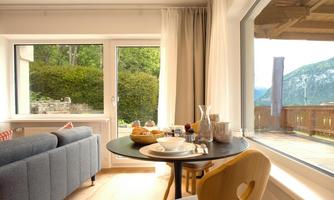 A cozy living space with a sofa and a table. Large windows offer a view of the garden and the mountains.