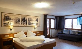 A modern hotel room with a double bed, a sofa, and large windows. Pictures of mountain landscapes hang on the wall.